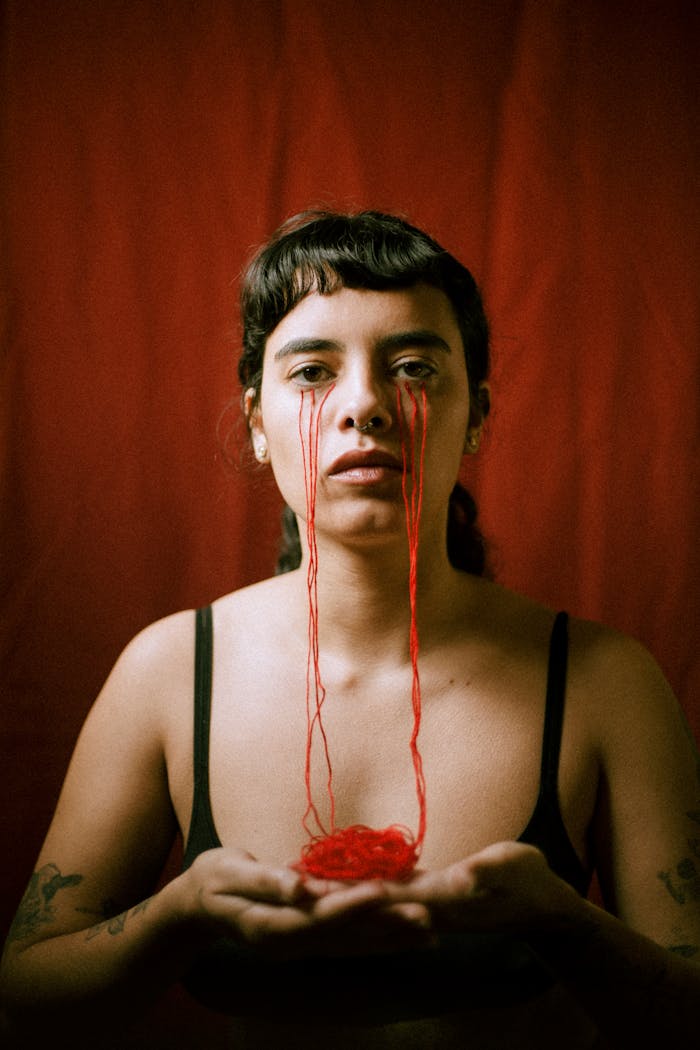 Artistic portrait of a woman with red threads mimicking tears, set against a red background.