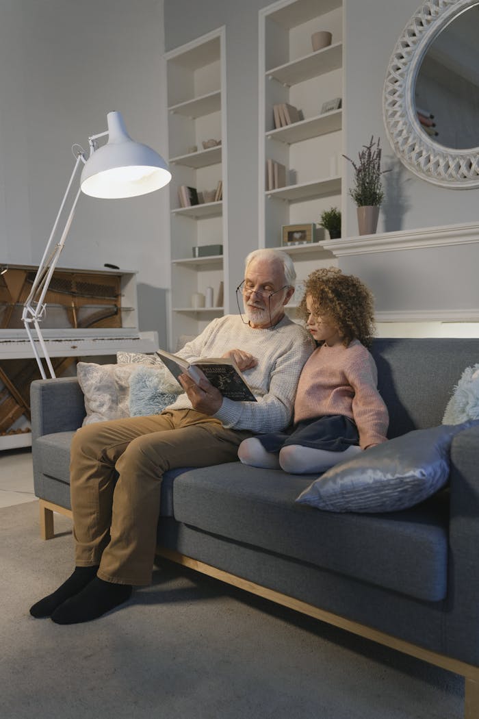 Elderly man and young girl reading together on a cozy sofa under a lamp.